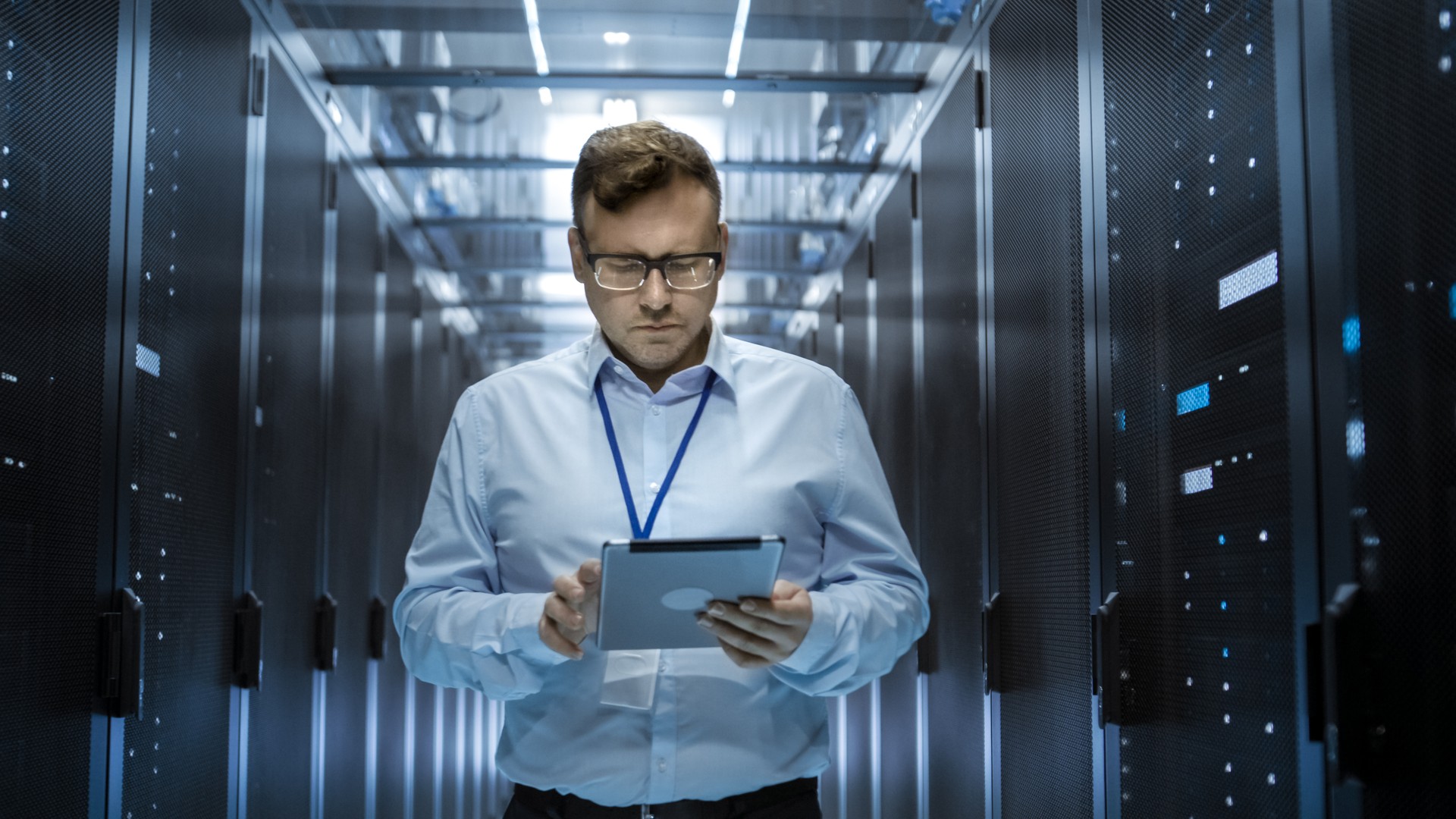 IT Technician Walks Through Rows of Server Racks in Data Center. Simultaneously He Works on a Tablet Computer.